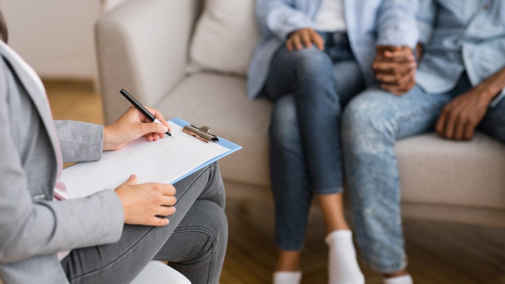 A therapist takes notes during a couples therapy session, with the couple holding hands on the couch in the background.
