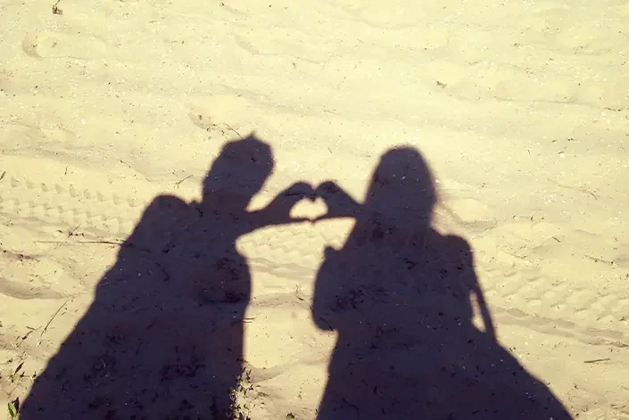 Shadows of a couple making a heart shape with their hands against a sandy background, symbolizing love and partnership.