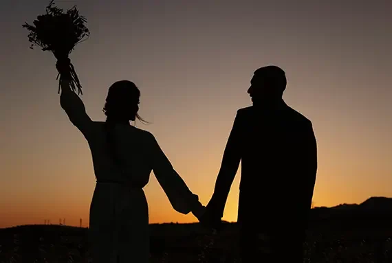 A silhouetted couple holding hands at sunset, with the woman holding a bouquet triumphantly in the air.