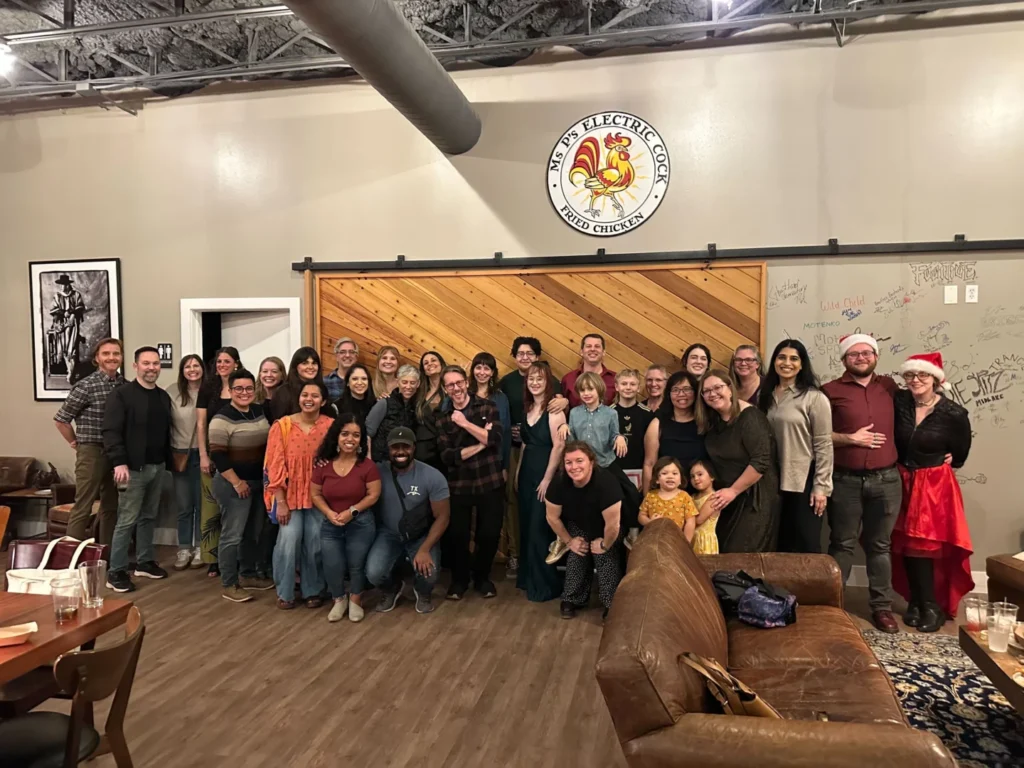 A large group of diverse coworkers posing for a holiday team photo in a rustic lounge setting with a wooden barn door.