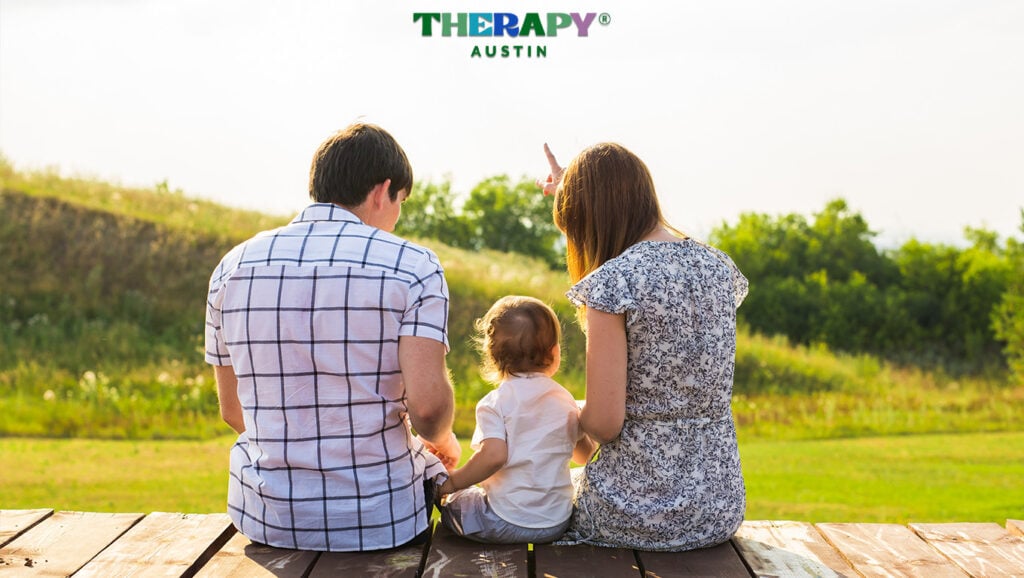 Parents and their young child sitting peacefully together looking toward a bright horizon, featuring the Therapy Austin logo.